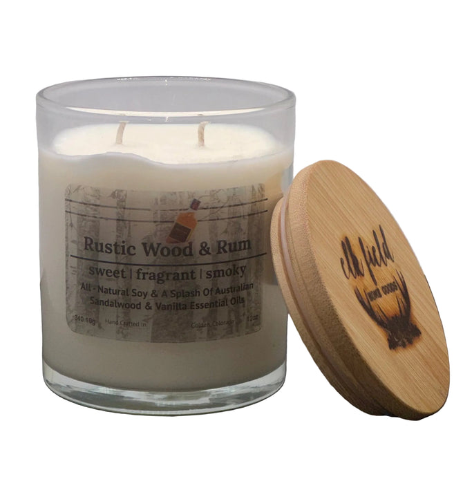 A two-wick candle in a clear glass jar with cream-colored wax, featuring a rustic paper label that reads 'Rustic Wood & Rum'. A round, natural bamboo lid with a dark burned logo sits next to the jar on a dark gray surface against a light gray background. Fall and Holiday Candle.